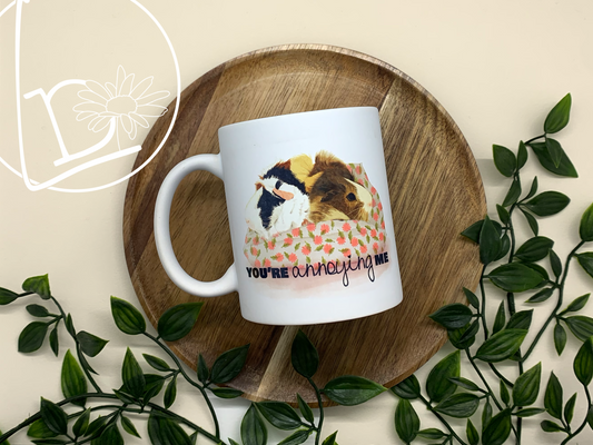 Guinea Pig Greeting Mug with an illustration of two guinea pigs, one blonde, one black, white and ginger. They sit facing away from each other on a white and pink cushion. The text says "You're annoying me""