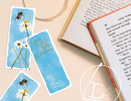 Bookmarks and books on a table. The bookmarks have a blue, watercolour sky background. The front of the bookmark has a bee about to land on a daisy. The back has the quote "love yourself the way bees love flowers."