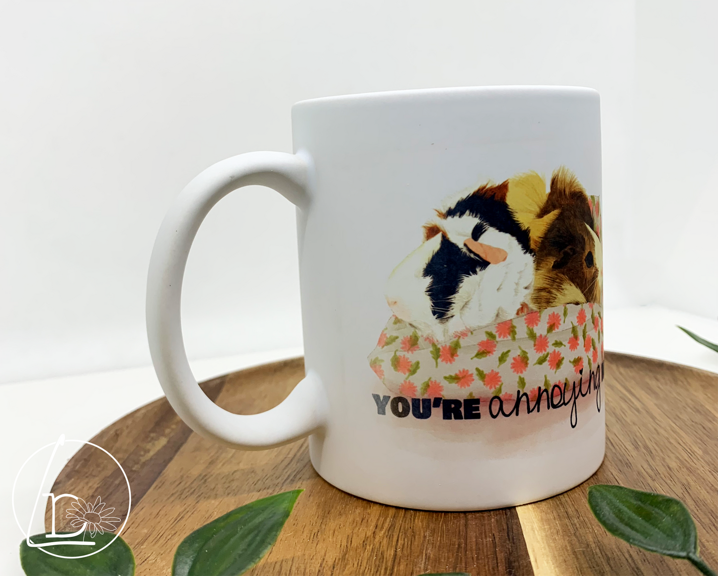 Guinea Pig Greeting Mug with an illustration of two guinea pigs, one blonde, one black, white and ginger. They sit facing away from each other on a white and pink cushion. The text says "You're annoying me""