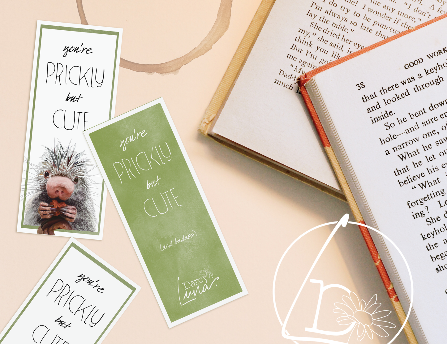 Bookmark and books on a table. The front of the bookmark has a green border and a prehensile tailed porcupine holding a heart. The slogan on both sides says "You're prickly but cute." The back has additional  text that says "and badass"