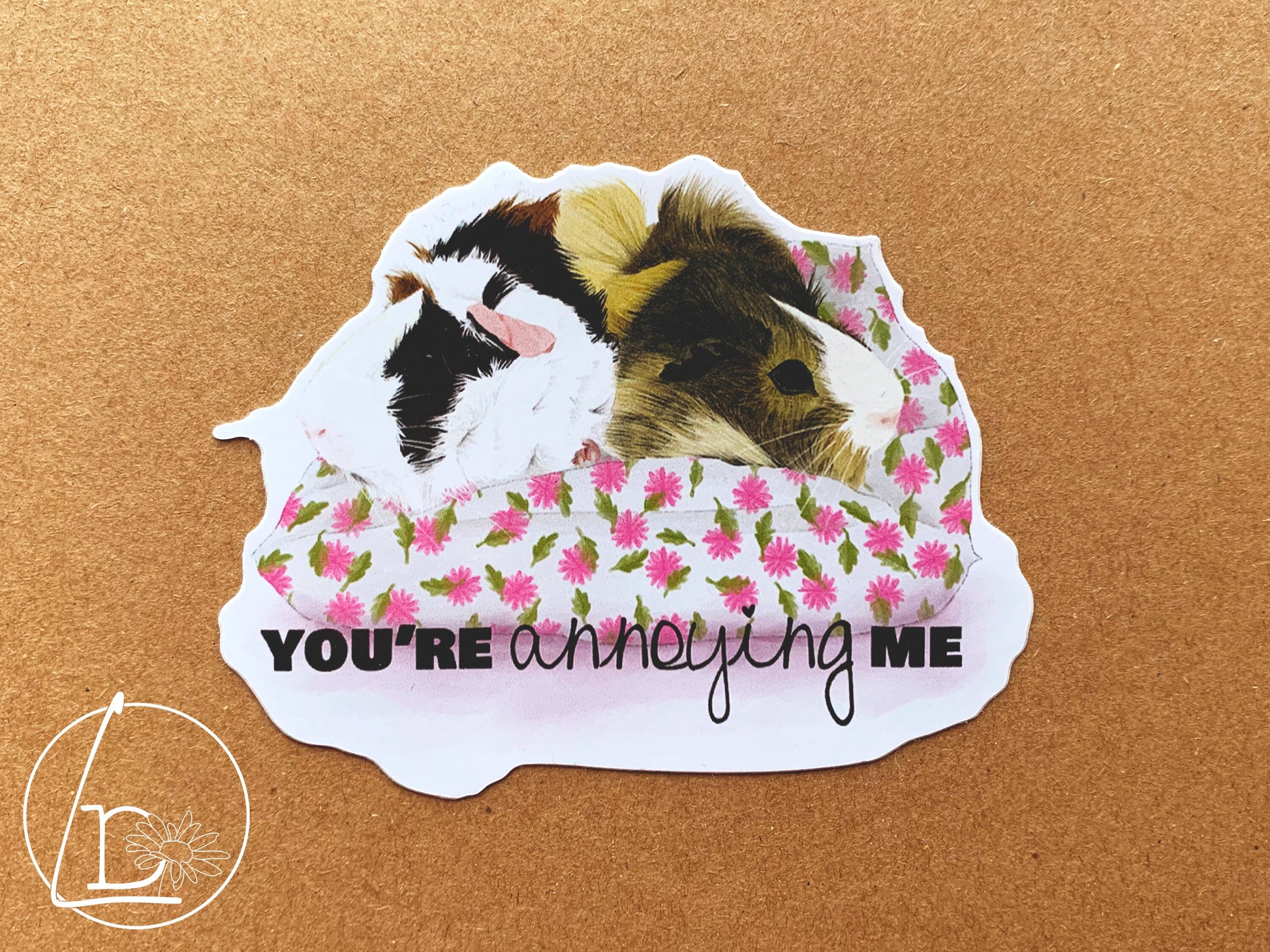 Guinea Pig Greeting Sticker with an illustration of two guinea pigs, one blonde, one black, white and ginger. They sit facing away from each other on a white and pink cushion. The text says "You're annoying me""