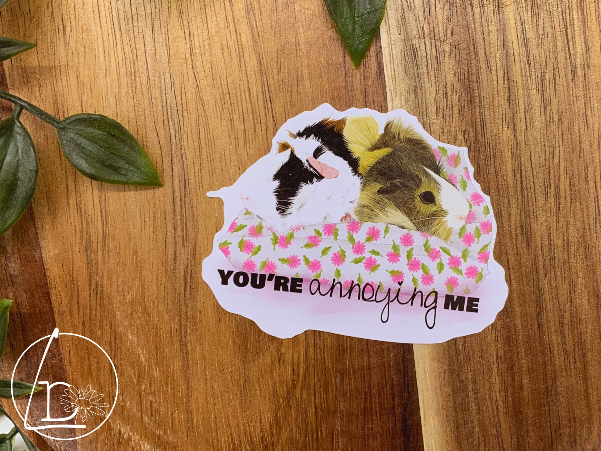 Guinea Pig Greeting Sticker with an illustration of two guinea pigs, one blonde, one black, white and ginger. They sit facing away from each other on a white and pink cushion. The text says "You're annoying me""