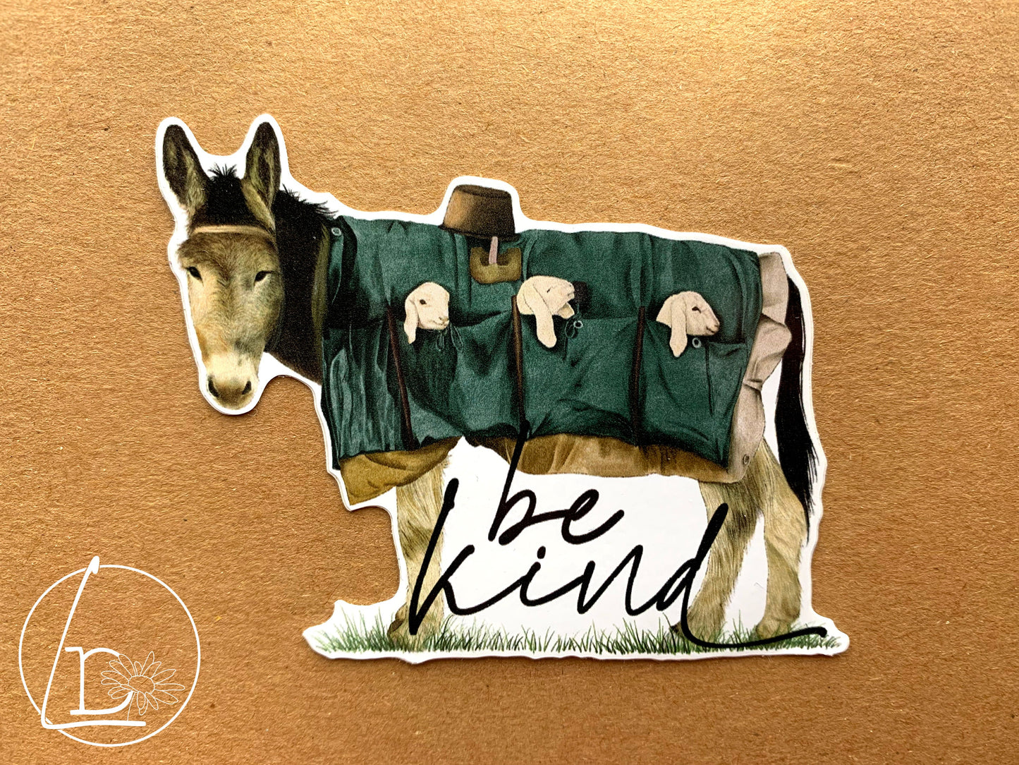 Illustrated sticker of a donkey carrying 3 lambs the pockets of his coat. The slogan says "be kind"