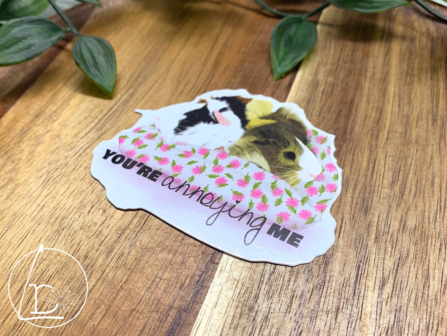 Guinea Pig Greeting Sticker with an illustration of two guinea pigs, one blonde, one black, white and ginger. They sit facing away from each other on a white and pink cushion. The text says "You're annoying me""