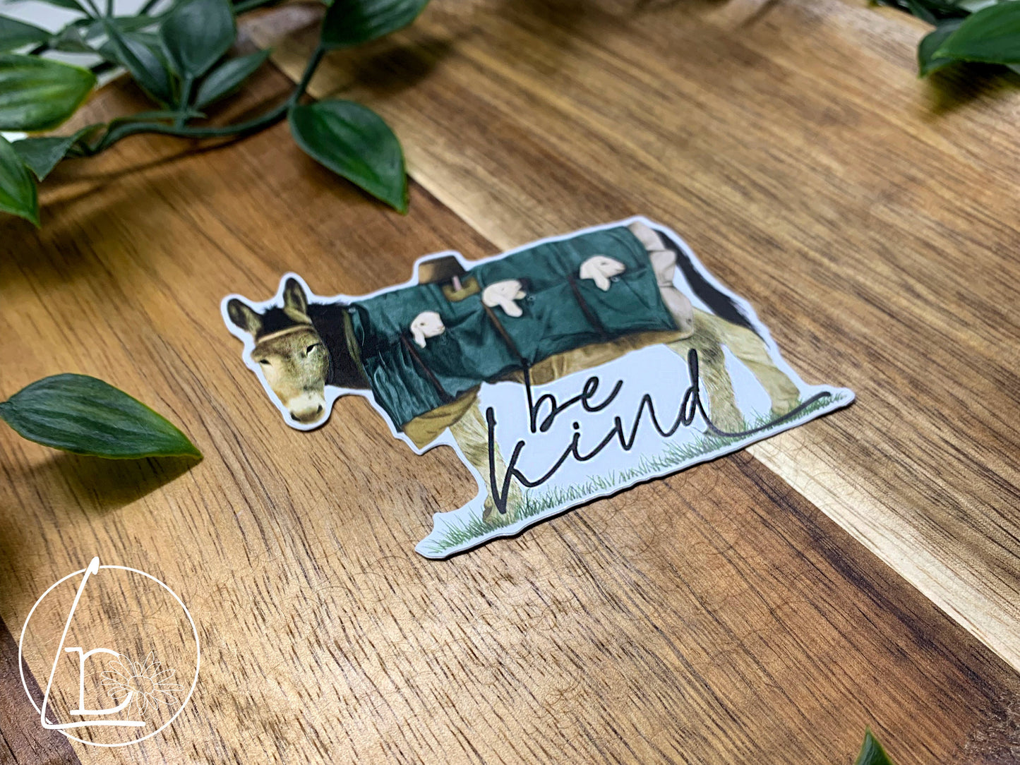 Illustrated sticker of a donkey carrying 3 lambs the pockets of his coat. The slogan says "be kind"