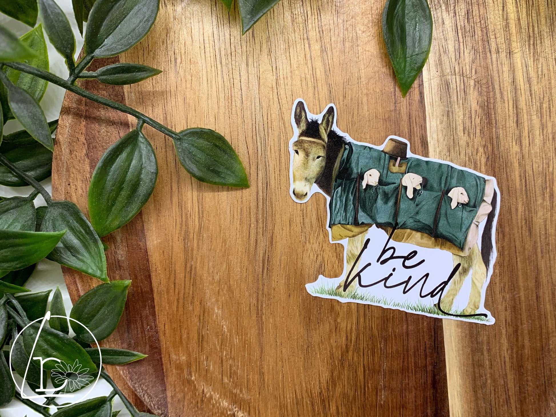 Illustrated sticker of a donkey carrying 3 lambs the pockets of his coat. The slogan says "be kind"